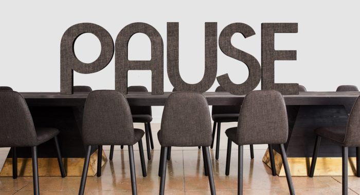 Mehrere leere graue Stühle stehen vor einem dunklen Konferenztisch. Auf dem Tisch steht in großen grauen Buchstaben aufrecht: PAUSE.