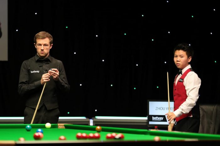 Viertelfinale der UK Championship: Jack Lisowski steht am Tisch, kreidet sein Queue und plant seine nächste Safety. Zhou steht daneben an seinem Platz und schaut gespannt auf den Tisch.