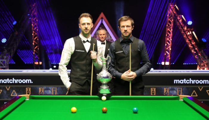 Judd Trump und Jack Lisowski posieren vor dem Finale der World Grand Prix mit der Trophäe. Im Hintergrund steht Referee Brendan Moore.