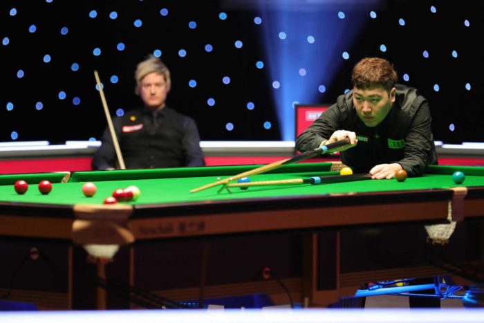 Yan Bingtao beim Lochen mit dem Hilfsqueue. Im Hintergrund sitzt Neil Robertson auf seinem Stuhl und kann nur zuschauen.