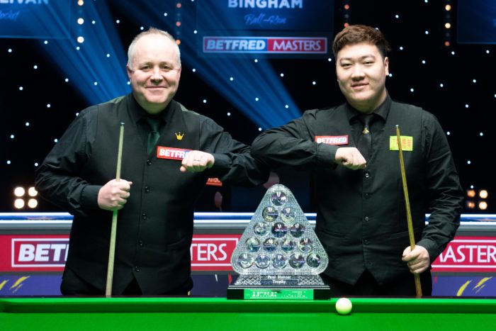 Vor Beginn des Masters Finals geben sich John Higgins und Yan Bingtao einen freundlichen Ellenbogencheck und lächeln in die Kamera. Vor ihnen steht die "Paul Hunter Trophy": Die Kristalltrophäe mit den 15 Bällen, wie die Roten beim Anstoß im Dreieck angeordnet sind.
