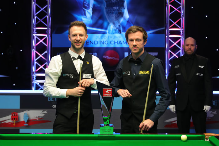 Judd Trump und Jack Lisowski geben sich vor dem Finale den in dieser Saison üblichen Ellenbogen-Check und lächeln in die Kamera. Im Vordergrund steht die Gibraltar Open Trophäe und im Hintergrund der Referee Colin Humphries.