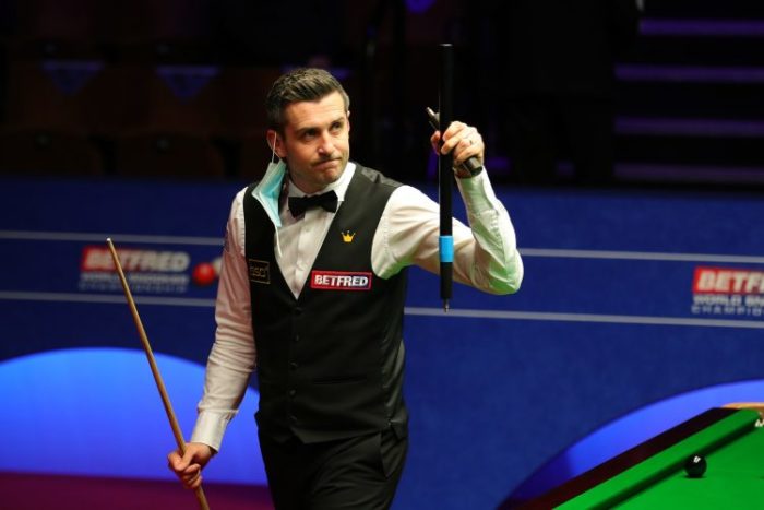 Nach seinem Viertelfinale verlässt Mark Selby die Arena. Er winkt mit seiner Queue-Verlängerung ins Publikum und hat seinen Mund-Nasen-Schutz schon überm Ohr hängen.