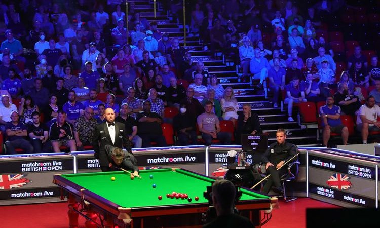 Das Match zwischen Mark Selby und Ali Carter bei den British Open. Selby ist am Tisch und im Stoß, Ali Carter sitzt in der Ecke auf seinem Stuhl. Der Hauptfokus der Kamera liegt jedoch auf den gefüllten Zuschauerrängen im Hintergrund.