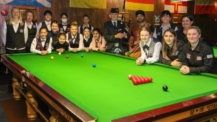 Die 16 Spielerinnen des Achtelfinals inklusive der Finalistinnen Reanne Evans und Mink Nutcharut haben sich für ein Gruppenfoto mit einem Dudelsackspieler und einem Trommler um einen Snookertisch versammelt.
