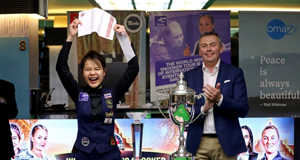 Weltmeisterin Baipat hält die Tourkarte hoch. Sie lacht. Neben ihr steht Jason Ferguson, vor ihr der WM-Pokal.