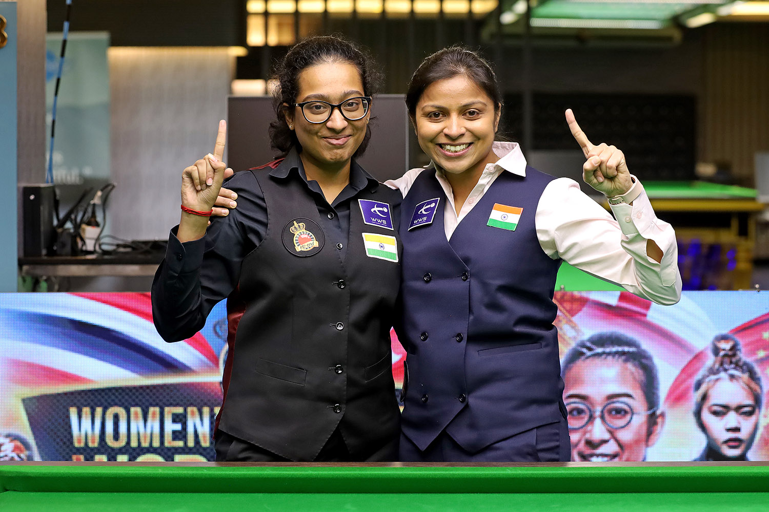 Amee Kamani und Anupama-Ramachandran stehen Arm in Arm an einem Snookertisch und halten einen Zeigefinger hoch.