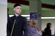 Liam Graham Graham, ein junger Mann mit dunklen Haaren steht in Standard-Snookerbekleidung mit dem Queue in der Hand da und wirft einen teils zufrieden teils beobachtenden Blick zur Seite. Im Hintergrund sehen Zuschauende in eine andere Richtung.