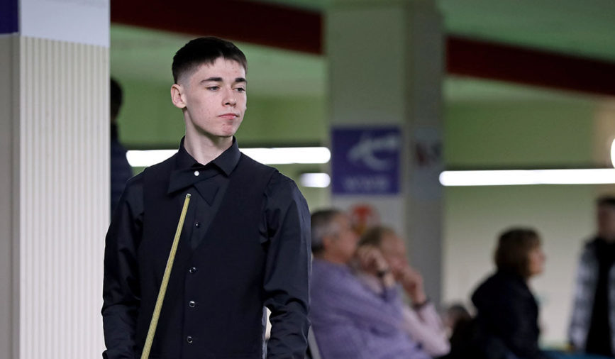 Liam Graham Graham, ein junger Mann mit dunklen Haaren steht in Standard-Snookerbekleidung mit dem Queue in der Hand da und wirft einen teils zufrieden teils beobachtenden Blick zur Seite. Im Hintergrund sehen Zuschauende in eine andere Richtung.