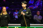 Si Jiahui, ein junger etwas schlacksiger chinesischer Spieler steht überlegend am Snookertisch und kreidet sein Queue. Er trägt schwarze Weste mit Hemd und dazu eine knallblaue Fliege.