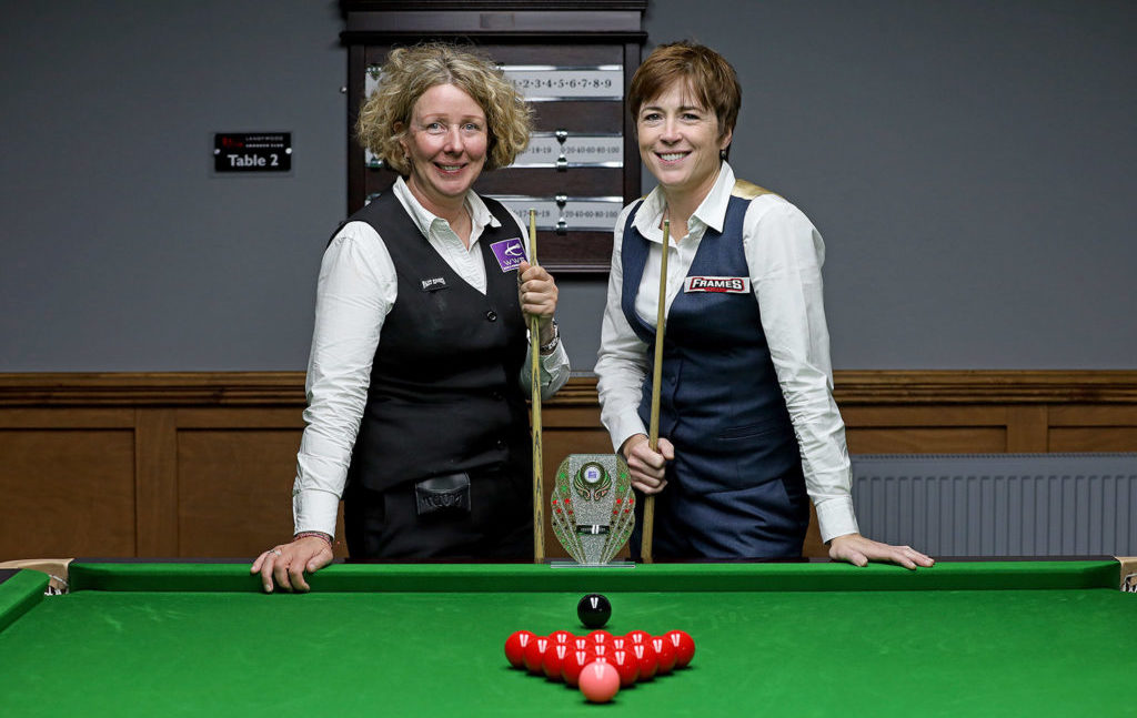 Zwei Frauen mittleren Alters stehen an einem Snookertisch und lächeln in die Kamera.