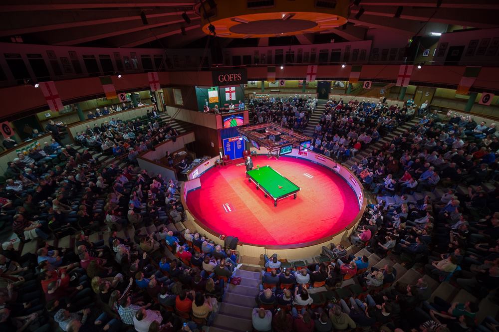 Ein Snookertisch steht in einer Manege auf rotem Teppich. Ringsherum steigen kreisförmig die gefüllten Zuschauerränge an, die auf den Start des Turniers warten.