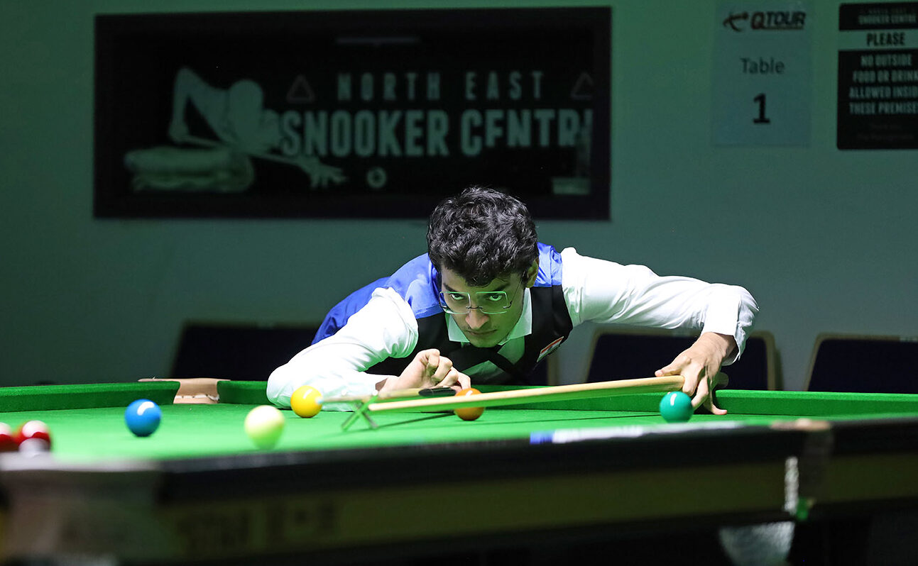 Ein Spieler mit sehr vollem dunklen Haar und randloser Snookerbrille mit hohen Gläsern visiert einen Stoß mit dem Hilfsqueue an. Sein Blick ist fokussiert und wirkt dabei fast etwas wütend.