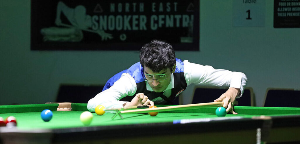 Ein Spieler mit sehr vollem dunklen Haar und randloser Snookerbrille mit hohen Gläsern visiert einen Stoß mit dem Hilfsqueue an. Sein Blick ist fokussiert und wirkt dabei fast etwas wütend.