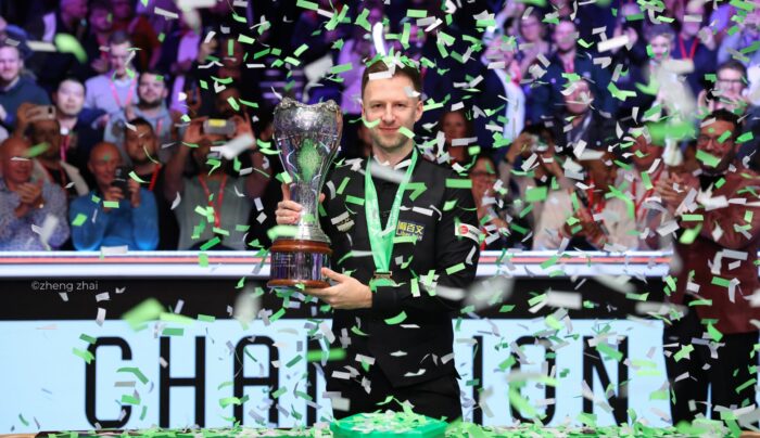 Judd Trump hält lächelnd die Trophäe der UK Championship hoch, während er im Konfetti Regen steht. Auf der Bande hinter ihm steht "Champion".