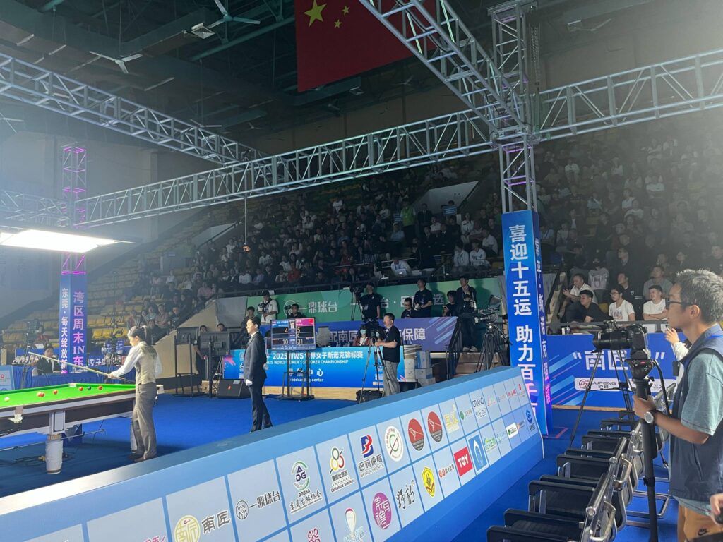 Eine Halle mit einem Snookertisch. Es ist eine Werbebande zu sehen, eine volle Tribüne.
