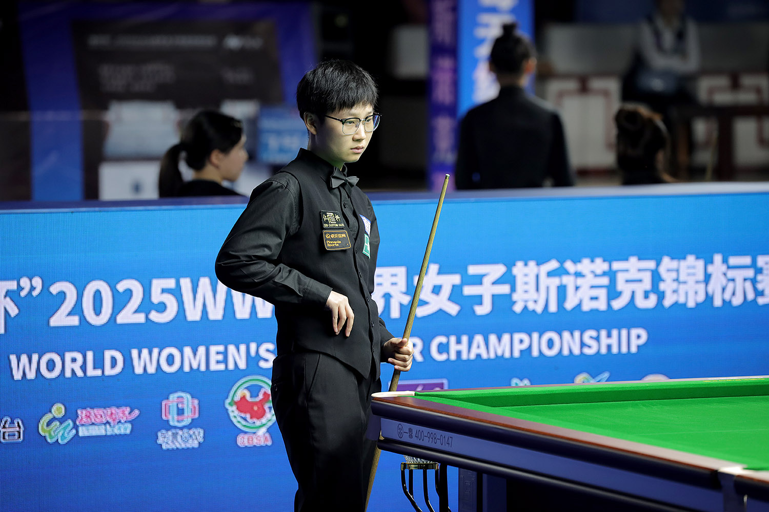 Eine junge chinesische Spielerin mit kurzen Haaren und Brille steht mit dem Queue in der Hand am Snookertisch und schaut sich die Lage der Bälle an.