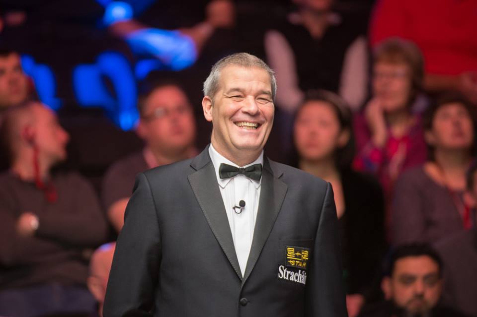 Jan Verhaas in mit Fliege steht lachend in einer Arena mit Publikum im Hintergrund.