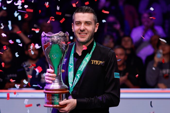 Mark Selby hält lächelnd die Trophäe der UK Championship, während Konfetti auf ihn herunterregnet.