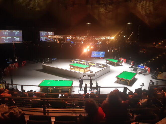 Blick von den Zuschauerrängen auf die Tische im Tempodrom. Nur noch am Tisch im Vordergrund wird gespielt. Xu Si, Mark Selby und Schiedsrichterin Anastasiya Tuzikova stehen alle an der Fußbande. Die meisten Roten sind noch auf dem Tisch.