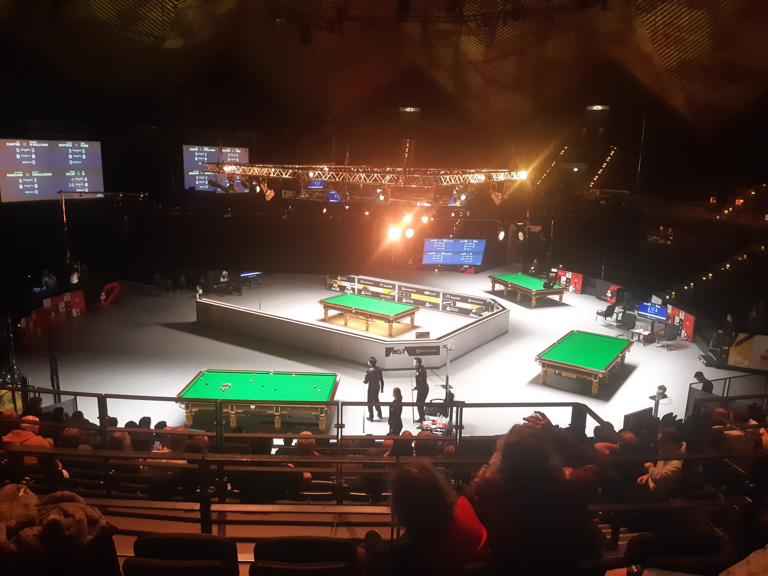 Blick von den Zuschauerrängen auf die Tische im Tempodrom. Nur noch am Tisch im Vordergrund wird gespielt. Xu Si, Mark Selby und Schiedsrichterin Anastasiya Tuzikova stehen alle an der Fußbande. Die meisten Roten sind noch auf dem Tisch.