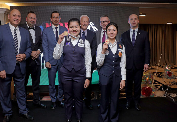 Panchaya Channoi und Jel Narucha halten strahlend ihre WSF Championship U-21 Medallien in die Kamera. Im Hintergrund postiert sich eine Riege wichtiger, in Anführungszeichen, Männer um Jason Ferguson, die die Bildkomposition zerstören.