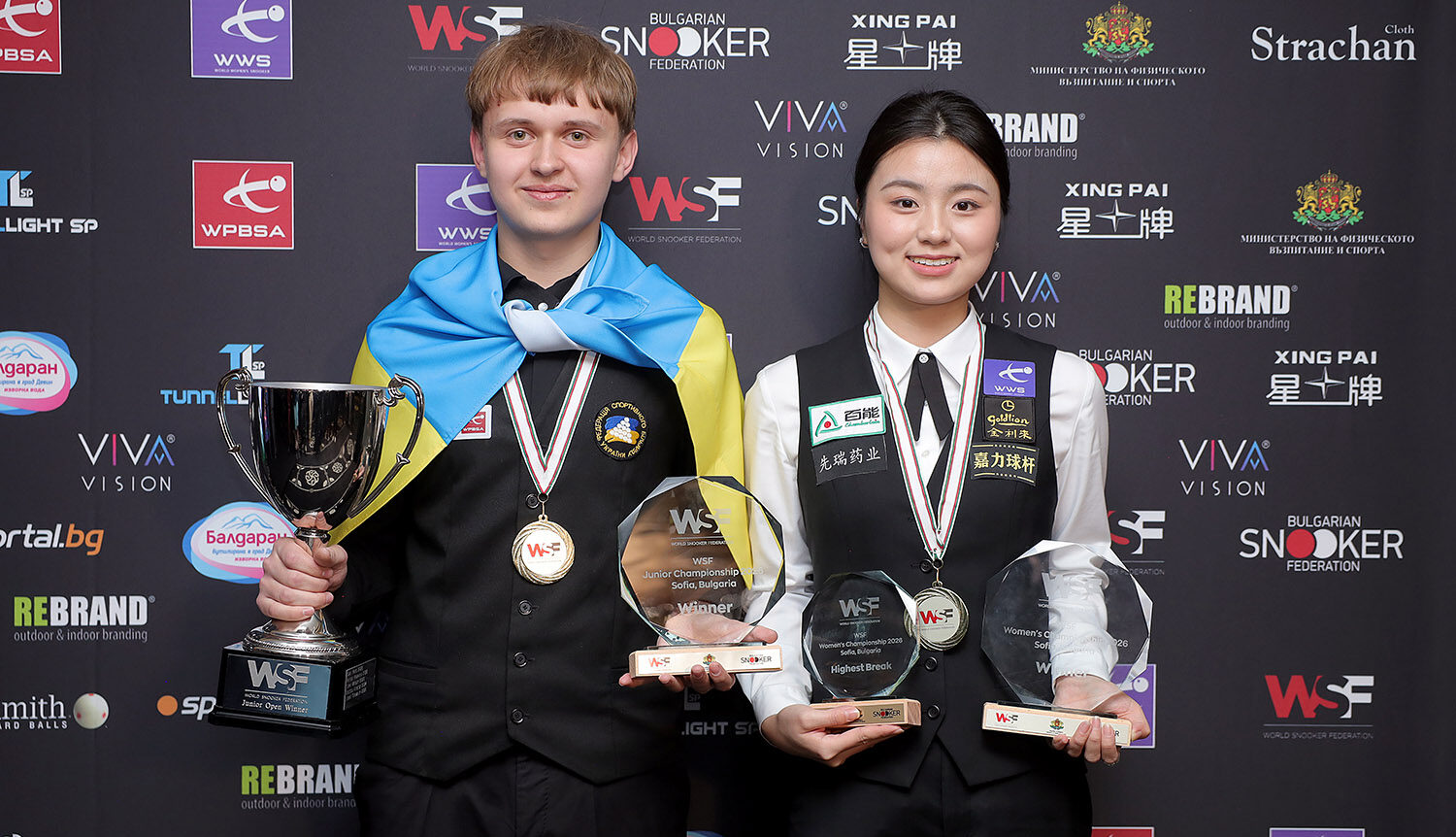Michael Larkov und Bai Yulu posieren mit ihren Trophäen. Links steht Larkov, mit einer Ukraine-Flagge um den Hals, der Silbertrophäe der WSF Junior Championship in der rechten und einer rundlichen Glastrophäe in der linken Hand. Rechts neben ihm steht bei Yulu, sie hält zwei ähnliche Glastrophäen für den Titel und das höchste Turnierbreak.