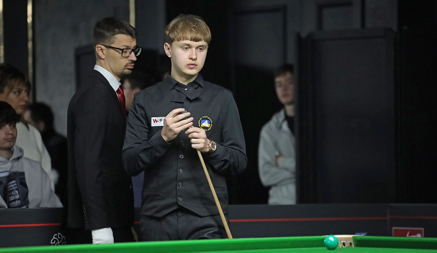 Ein junger Mann kreidet sein Queue und blickt nachdenklich auf den Snookertisch. Seine dunkelblonden kurzen Haare sind gerade auf der Stirn geschnitten.