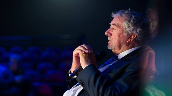 John Virgo im Profil bei einem Seniors-Event. Er sitzt ruhig und hat die Hände vor der Brust gefaltet.