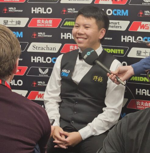 Zhou Yuelong sitzt mit gefalteten Händen vor der Werbebannertafel und strahlt glücklich über beide Backen. Ihm wird ein Mikrofon ins Gesicht gehalten und im Bildrand erahnt man die ihn interviewenden Journalisten.