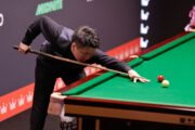 Zhou Yuelong ist im Stoß, er spielt ein Break um den Spot der Schwarzen herum. Im Hintergrund sieht man die Trennwand, die die beiden Tische bei der Weltmeisterschaft im Crucible trennt.