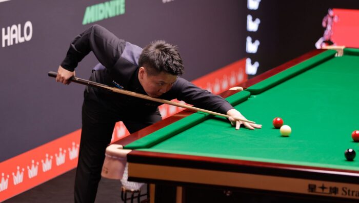 Zhou Yuelong ist im Stoß, er spielt ein Break um den Spot der Schwarzen herum. Im Hintergrund sieht man die Trennwand, die die beiden Tische bei der Weltmeisterschaft im Crucible trennt.