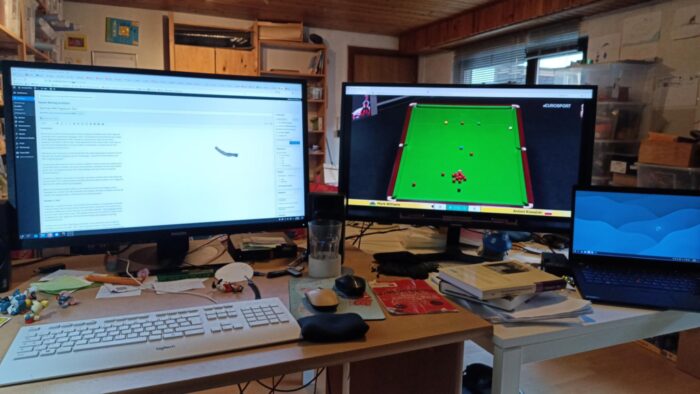 ein unaufgeräumtes Arbeitszimmer. ein Laptop, ein kleiner PC und zwei große Monitore füllen das Bild aus , vor dem Ganzen auf dem Schreibtisch eine Tastatur, ein Glas, Schlümpfe und diverser Krams. Auf dem linken Monitor der Text dieses WM Tagebuch s in einer Rohfassung , rechts das Match Mark Williams gegen Antoni Kowalski.