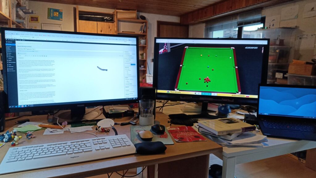 ein unaufgeräumtes Arbeitszimmer. ein Laptop, ein kleiner PC und zwei große Monitore füllen das Bild aus , vor dem Ganzen auf dem Schreibtisch eine Tastatur, ein Glas, Schlümpfe und diverser Krams. Auf dem linken Monitor der Text dieses WM Tagebuch s in einer Rohfassung , rechts das Match Mark Williams gegen Antoni Kowalski.