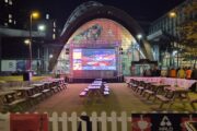 Public Viewing, leere Bänke, nachts. Die Leinwand zeigt noch das leere Crucible von innen.