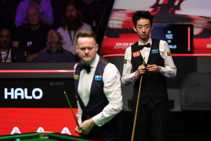 Shaun Murphy unscharf im Vordergrund am Tischrand. im Hintergrund Wu Yize, die Kamera fokussiert ihn.