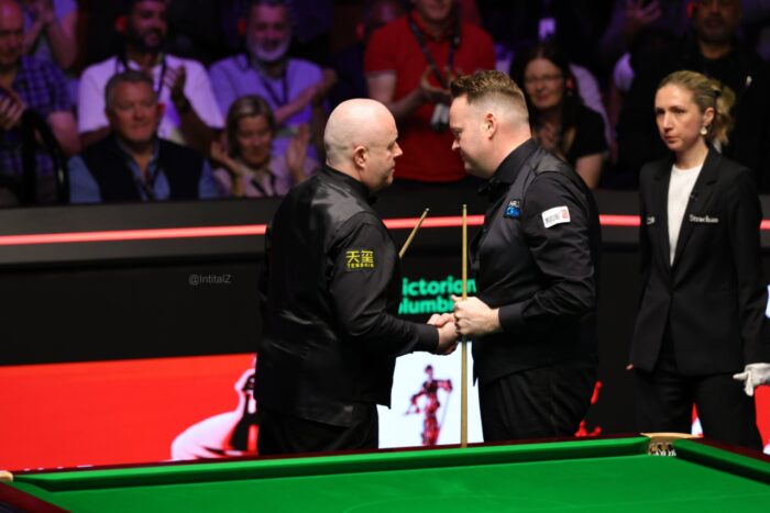 John Higgins und Shaun Murphy stehen hinter der Fußbande des Tisches im Crucible Theatre nach ihrem Halbfinale. Higgins schüttelt gratulierend die Hand und sagt etwas zu ihm. Im Hintergrund steht die Schiedsrichterin des Halbfinals Tatiana Woollaston.