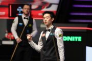 Wu Yize steht im Vordergrund und schaut nach vorne, er ballt die rechte Hand zur Faust. Im Hintergrund kommt sein Halbfinal-Gegner mit dem Queue zu ihm nach vorne gelaufen.