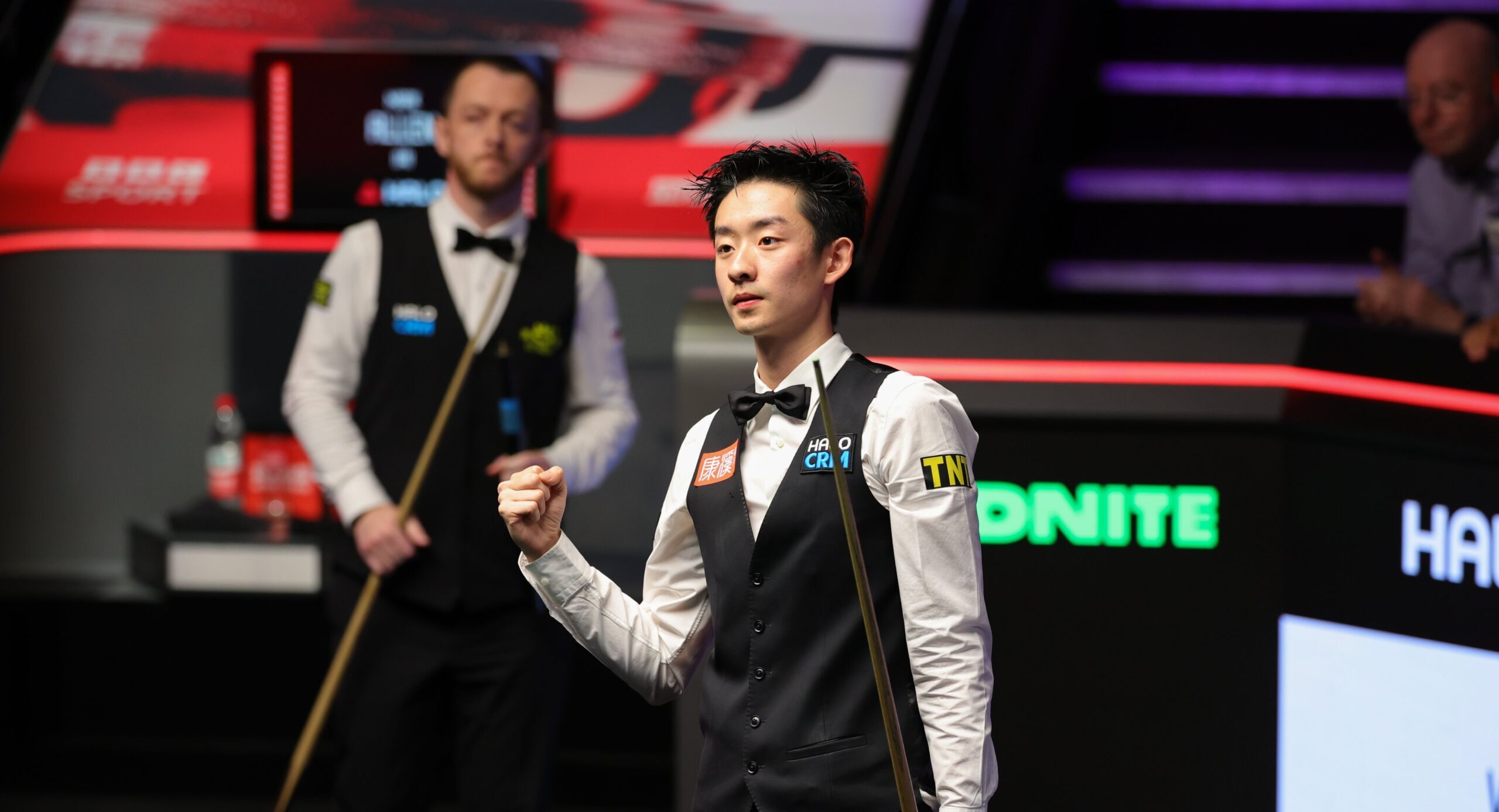 Wu Yize steht im Vordergrund und schaut nach vorne, er ballt die rechte Hand zur Faust. Im Hintergrund kommt sein Halbfinal-Gegner mit dem Queue zu ihm nach vorne gelaufen.
