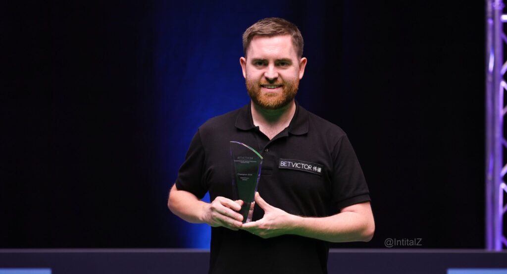Jak Jones hält die Trophäe der Championship League in der Hand und lächelt.